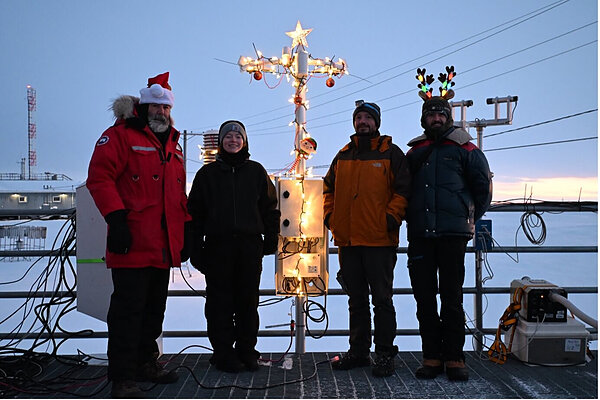 Four scientists in cold weather gear in the Arctic posing with an anemometer decorated with Christmas lights.